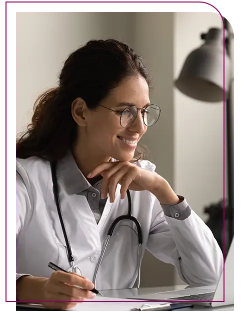 Mulher branca de oculos, cabelo preso, jaleco branco sentada em frente ao notebook