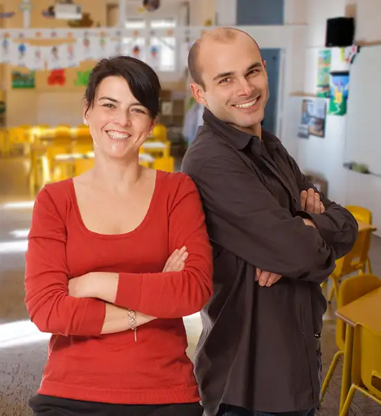 um homem e uma mulher professores de braços cruzados sorrindo em frente a uma sala de educação infantil