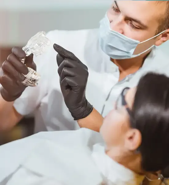 homem dentista de jaleco e máscara e luvas explicando algo para uma paciente que está com óculos de proteção em uma cadeira