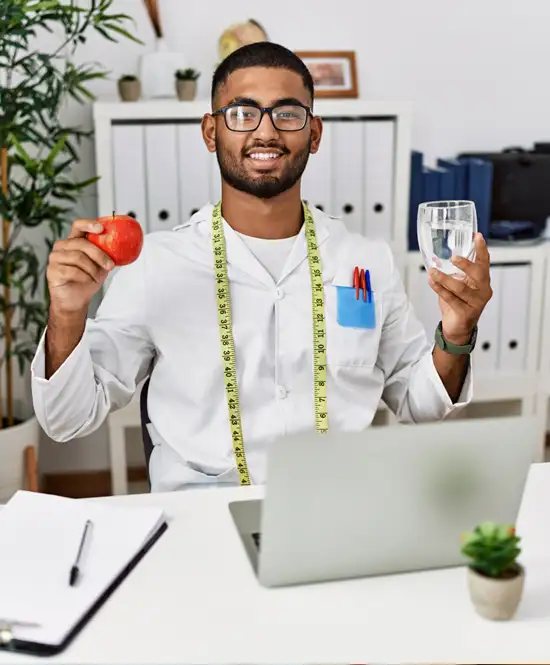 homem nutricionista sorrindo com jaleco branco fita métrica branca no pescoço um copo de água em uma mão e uma fruta em outra