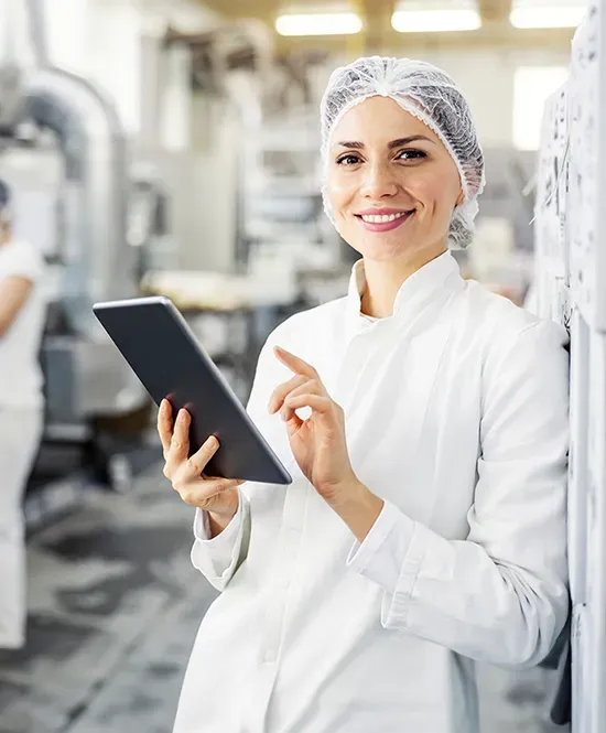 mulher com jaleco branco e touca nos cabelos segurando tablet e sorrindo em uma indústria