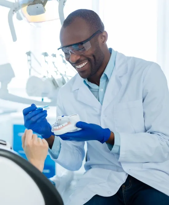 homem negro dentista sorrindo segurando prótese dentária com luvas e jaleco branco e óculos de proteção