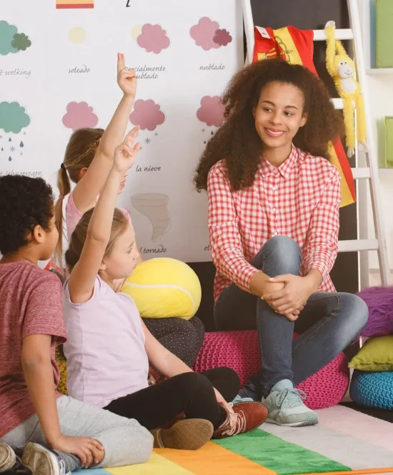 mulher professora sorrindo sentada no chão de uma sala de educação infantil com diversas crianças e algumas crianças levantando a mão