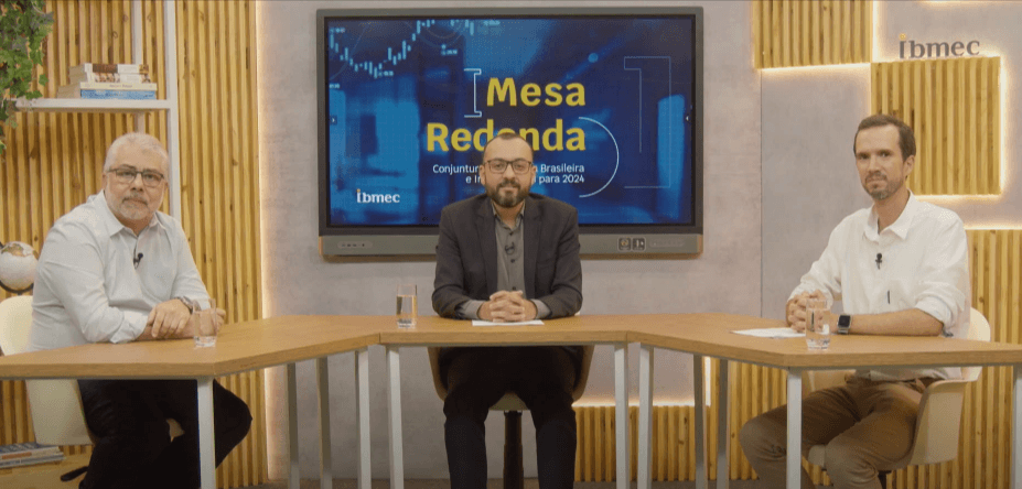 Três homens brancos sentados ao redor de uma mesa de madeira, com cenário que se assemelha a uma sala de estar e uma televisão atrás deles escrito Mesa Redonda