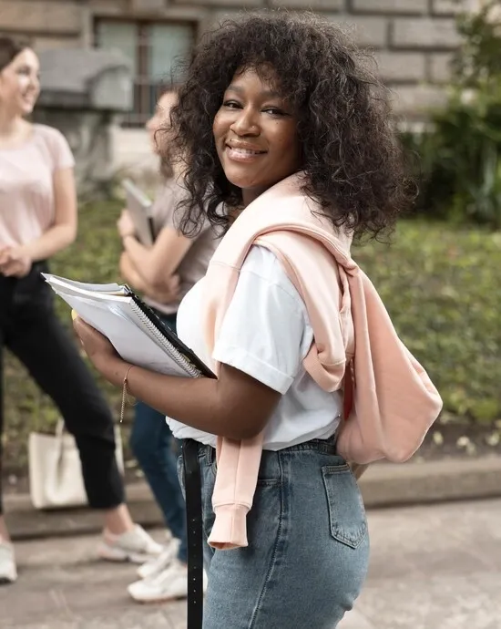 mulher negra de cabelos soltos sorrindo com mochila nas costas e cadernos nas mãos