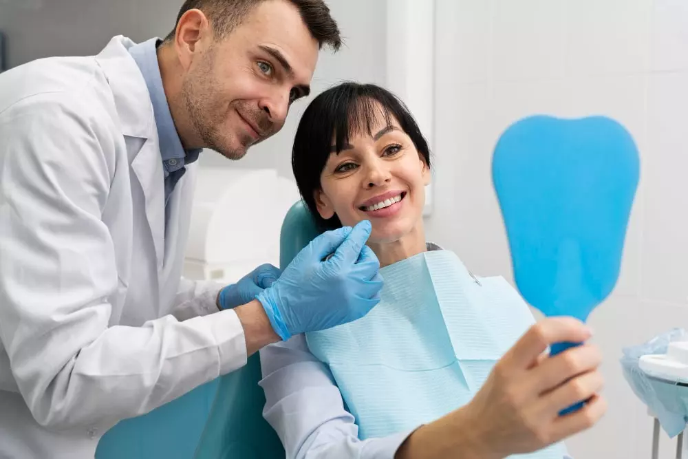 Dentista homem de jaleco branco usando luvas azul claro com dedo na boca de paciente mulher sorridente vendo num espelho de mão o resultado.