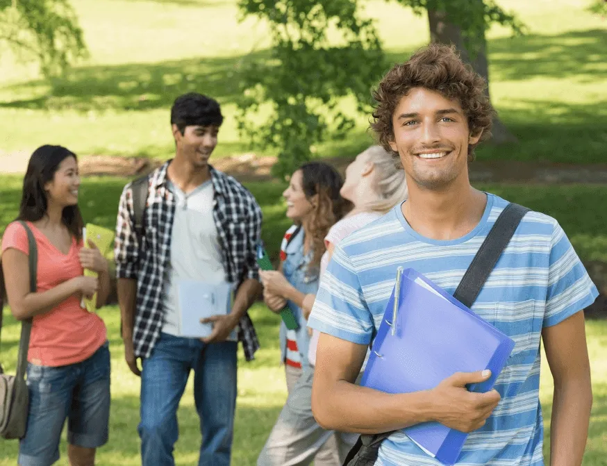 homem branco sorrindo com caderno na mão em frente a um jardim com outros estudantes conversando atrás