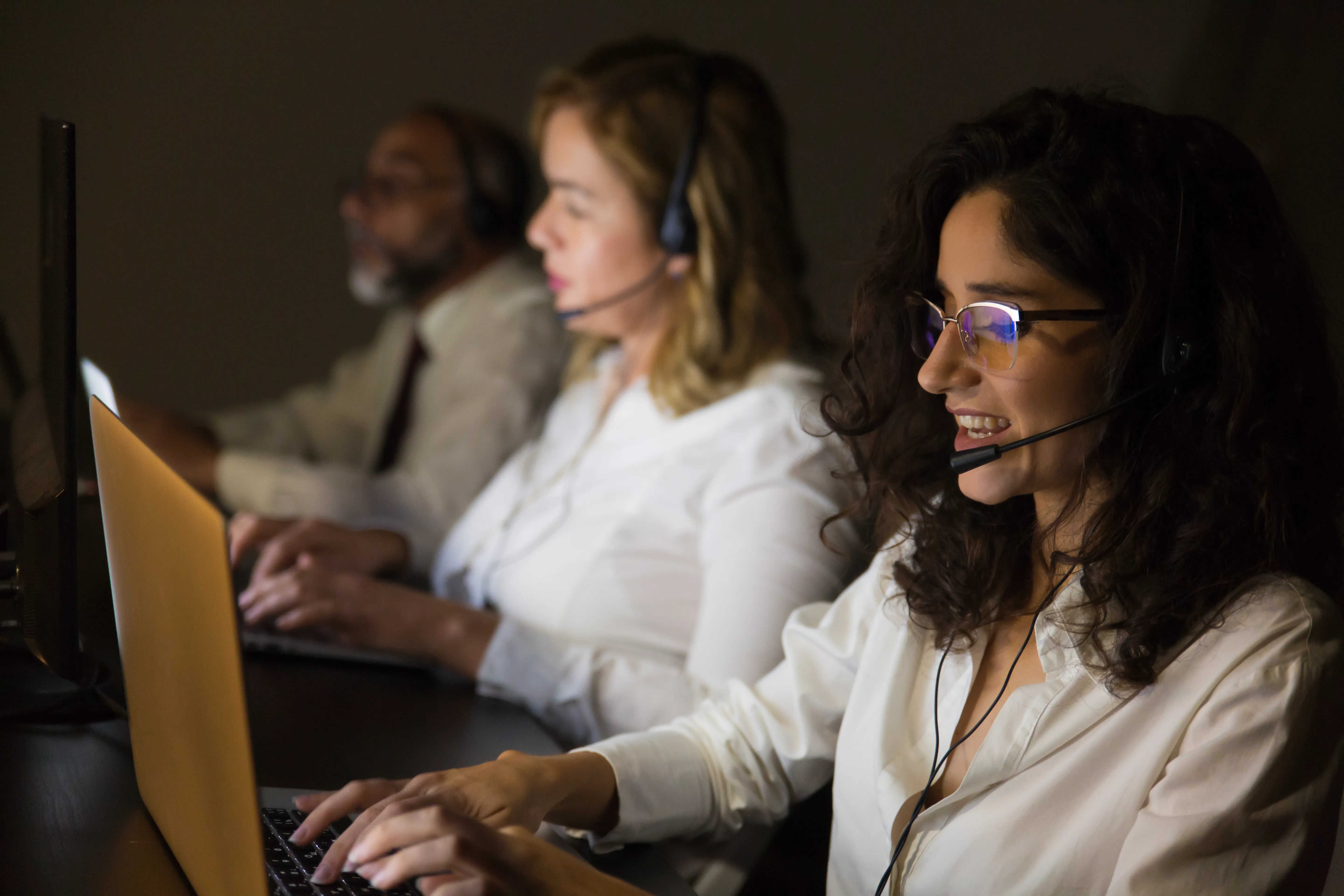 duas mulheres e um homem sentados a frente de uma mesa com computadores e headset